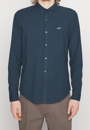 Muž v tmavě modré košili s dlouhým rukávem zapnuté na knoflíky, s malým bílým logem na hrudi a v světle hnědých kalhotách.