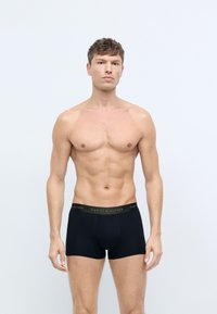 Sorte bomulds-boxershorts med en blød elastisk talje, der har "TOMMY HILFIGER" i guld-bogstaver. Modellen står med et neutralt udtryk.