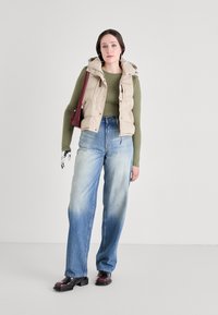 Beige puffer vest over a grønn langermet topp, sammen med lyseblå brede bukser. Sorte chunky sko og en rød veske fullfører antrekket.