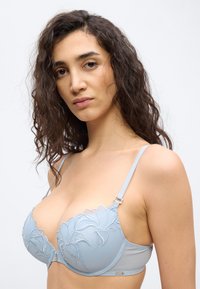 Žena s dlhými kučeravými tmavými vlasmi, ktorá nosí svetlomodrú podprsenku s kvetinovou výšivkou, pozerá sa priamo do kamery na jednoduchom pozadí.