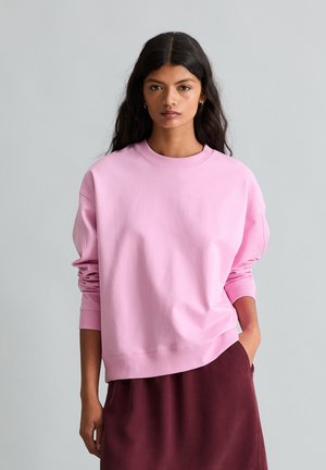 Kobieta z długimi ciemnymi włosami, ubrana w luźną różową bluzę i ciemnoczerwoną spódnicę, jedną ręką w kieszeni, o neutralnym wyrazie twarzy, na jednolitym szarym tle.