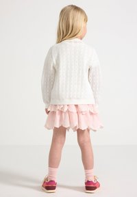 Maglione bianco lavorato a maglia con motivo testurizzato, gonna rosa a strati con volant, calzini rosa e scarpe colorate. Vista da dietro.