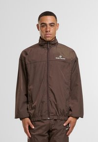 Mladenič s kratkimi lasmi in pegami, oblečen v rjavo zip-up jakno Sergio Tacchini in ujemajoče se hlače, stoji ob ozadju brez vzorca.