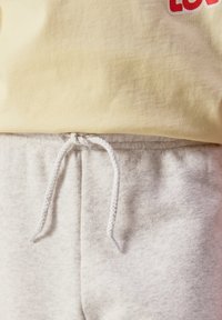 Gros plan sur un pantalon de survêtement beige avec un cordon blanc noué à la taille, associé à un haut jaune montrant partiellement un texte rouge.