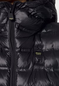 Černá proš�ívaná péřová bunda s lesklou texturou. Má přední zip a na hrudi žlutý štítek s nápisem "Blauer U.S.A.".