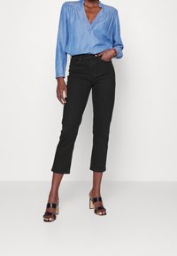 Camisa de manga larga en tono azul claro, con escote en forma de V y hombros fruncidos; jeans negros recortados; sandalias negras de tacón con tacones en bloque.