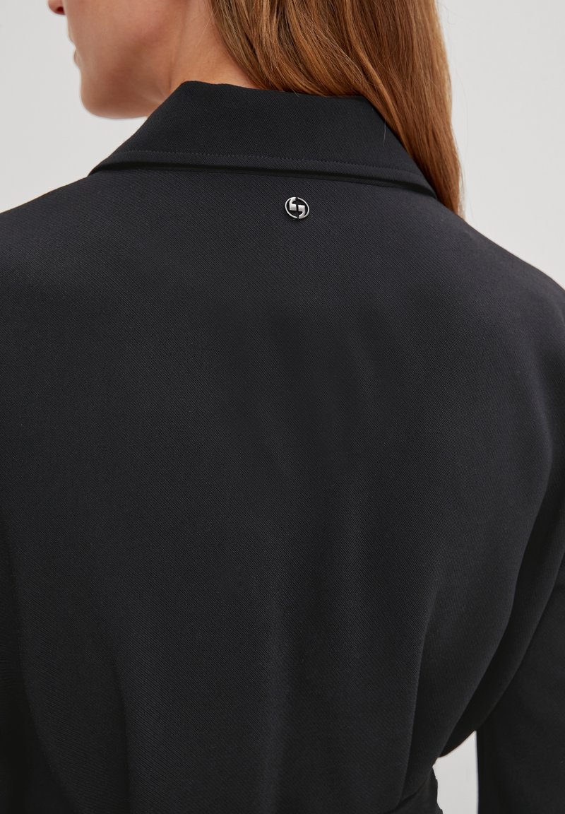 Femme aux cheveux châtain clair portant un vêtement noir à col, montrant un petit logo rond en argent sur le haut du dos.