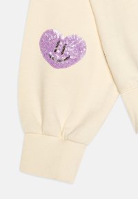 Tessuto color crema con un polsino a costine caratterizzato da un design a cuore di paillettes viola, arricchito da dettagli neri di sorriso e occhi.