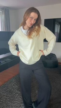 Krämfärgad stickad tröja med V-ringning, kombinerad med lösa svarta byxor. Bakgrunden visar en platt-TV och en mörk sittsäck.