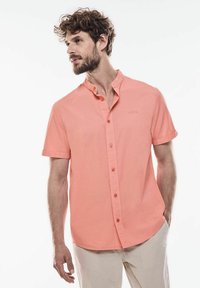 Krátký rukáv košile na knoflíky v korálové barvě, s mandarin límcem a diskrétním logem. Hladká látka s uvolněným střihem.