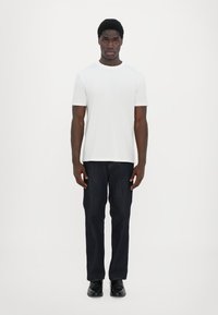 Hvit t-skjorte med korte ermer, blå denimjeans, svarte sko, står mot en ensfarget hvit bakgrunn. Enkel design, ingen synlige mønstre eller logoer.
