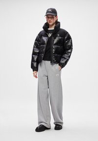 Mīksts melns jaku ar spīdošu virsmu, pelēks sporta krekls, brīvi pelēki sporta bikses un melnas kurpes, ko papildina melna cepure ar logo.