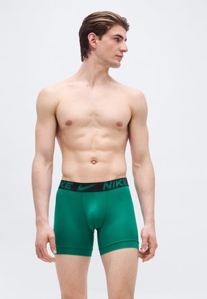 Man zonder shirt met kort bruin haar, gekleed in groene Nike boxershorts, staand en naar zijn linkerzijde kijkend tegen een effen lichte achtergrond.