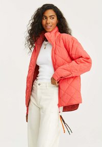 Nabity koralowy żakiet z wysokim kołnierzem i zamkiem błyskawicznym, o luźnym kroju, zestawiony z białą koszulą i jasnymi spodniami.