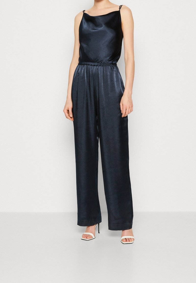 Femme portant une combinaison sans manches en satin noir avec des jambes larges et des sandales blanches à talons hauts, debout devant un fond uni.