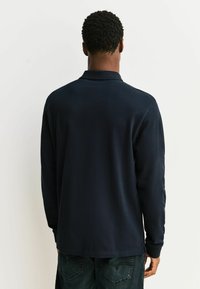 Polo de manga larga azul marino con cuello y puños acanalados, tela suave y aberturas laterales en el dobladillo. Combinado con vaqueros oscuros.