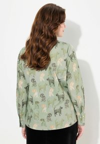 Camisa verde con un estampado de perro juguetón en negro, naranja y verde claro. Cuenta con mangas largas y puños con botones, con una textura suave de algodón.
