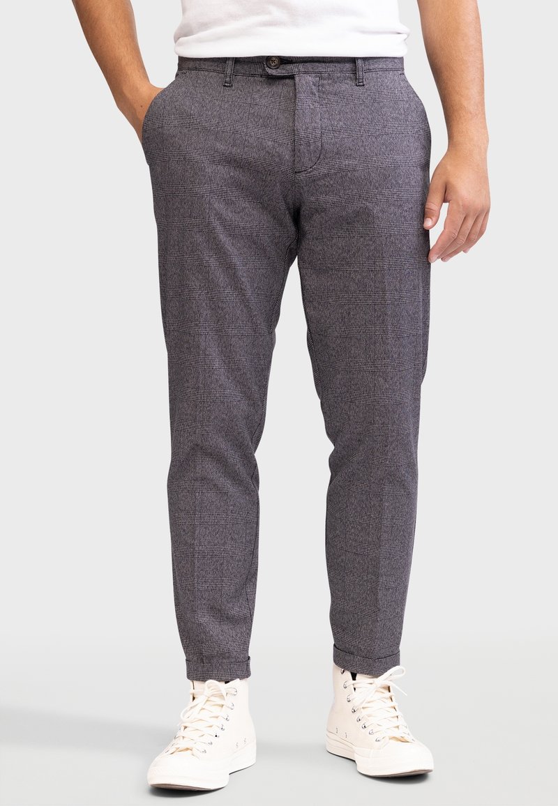 Homme portant un pantalon slim à carreaux gris et des baskets montantes blanches avec une chemise blanche rentrée.