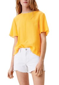 Top giallo a maniche corte con finitura testurizzata e orli ondulati, abbinato a pantaloni corti in denim bianchi con orlo sfrangiato.