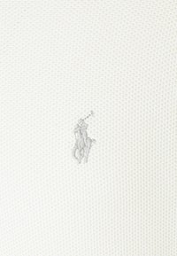 Tissu blanc texturé avec petit logo gris brodé représentant un joueur de polo à cheval maniant un maillet.