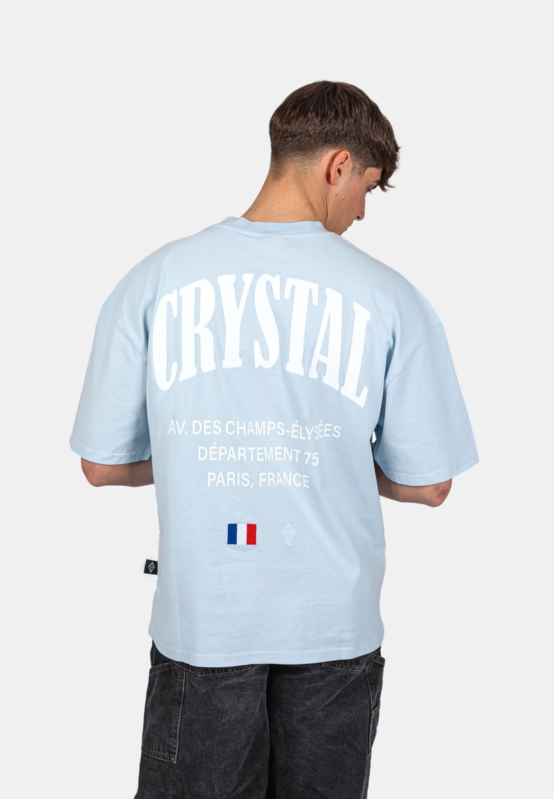 Světle modré oversized tričko má na zádech bílý potisk, který zahrnuje slovo "CRYSTAL" a malý detail francouzské vlajky.