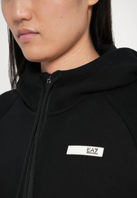Čierna mikina s kapucňou na zips, s hladkou textúrou a bielym logom EA7 na obdĺžnikovom štítku pri zipsovom uzávere.