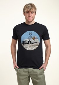 Černé bavlněné tričko s kulatým grafickým motivem vintage bílé auto s osobou vyklánějící se ven, umístěným na texturovaném modrém pozadí.