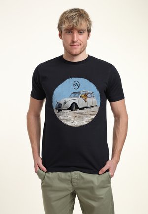 Černé bavlněné tričko s kulatým grafickým motivem vintage bílé auto s osobou vyklánějící se ven, umístěným na texturovaném modrém pozadí.
