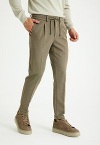 Beige Hose aus strukturiertem Stoff, mit gefaltetem Vorderteil, Kordelzugbund, schmal zulaufendem Bein und Seitentaschen, kombiniert mit braunen Sneakern.