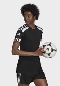 Schwarzes Sportshirt mit weißen Streifen auf den Schultern, kombiniert mit schwarzen Shorts. Hält einen schwarz-weißen Fußball mit Mustern.