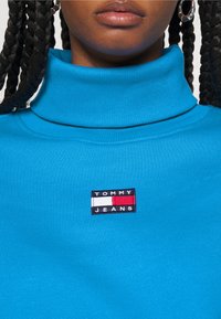 Niebieski bawełniany golf z fakturą ribbed, z prostokątną naszywką logo Tommy Jeans w kolorze czarnym, białym i czerwonym na klatce piersiowej.