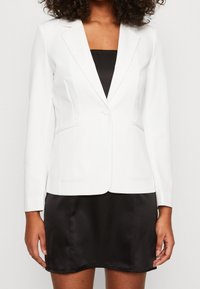 Blazer blanc à coupe ajustée, fermeture à un bouton, revers crantés et deux poches latérales, porté sur une robe en satin noire.