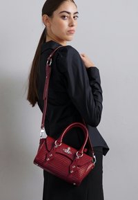 Bolsa de mão texturizada em croco vermelho com ferragens prateadas, alças duplas e uma alça ajustável, exibida contra uma parede neutra.