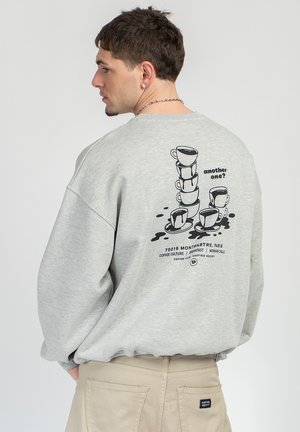 Hombre con sudadera gris clara con un gráfico negro de tazas de café apiladas y texto "¿Otra más?" además de detalles de un café de París en la parte trasera.