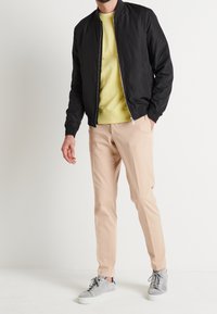 Svart bomberjacka med dragkedja fram, ljusgul sweatshirt under, beigefärgade byxor och grå sneakers med vita sulor.