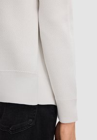 Langärmliges, hellgraues, strukturiertes Strickoberteil mit geripptem Saum und Bündchen, kombiniert mit dunklen Jeans, die kontrastierende Materialien betonen.