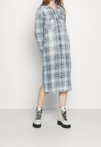 Długa koszulowa sukienka w niebiesko-biały prążek, z zapięciem na guziki z przodu, dwoma kieszeniami na piersiach oraz bocznymi rozcięciami, zestawiona z białymi butami na sznurówki.