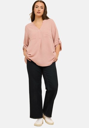 Vrouw draagt een lichtroze losse blouse met opgerolde 3/4 mouwen, zwarte wijde broek en witte sneakers, staand met handen in de zakken.