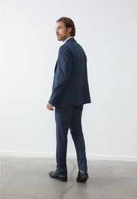 Marineblauw twee-delig pak van gestructureerde stof; op maat gemaakt snit met iets taps toelopende broek en zwarte nette schoenen. Minimale details.