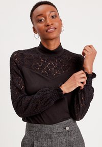 Haut noir avec col haut, doté de longues manches en dentelle et d'un tissu texturé. Le modèle porte un pantalon à carreaux gris.