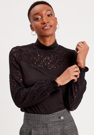 Haut noir avec col haut, doté de longues manches en dentelle et d'un tissu texturé. Le modèle porte un pantalon à carreaux gris.