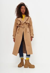 Trench coat beige con vita cin cin, bottoni doppi, e ampi risvolti, abbinato a jeans scuri e stivali marroni con lacci.