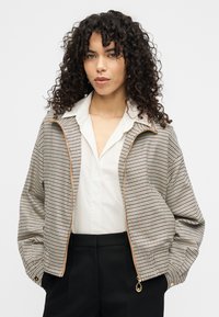 Καρό μπουφάν τύπου bomber με μπεζ και μαύρο σχέδιο, διαθέτει φερμουάρ μπροστά, γιακά και χρυσά μεταλλικά στοιχεία. Φορεμένο πάνω από λευκό πουκάμισο.