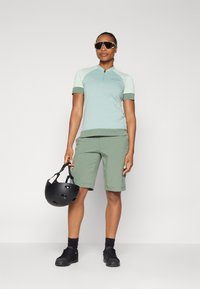 Top de manga corta, de color verde claro, con cuello de cremallera y acentos en verde más oscuro, combinado con pantalones cortos verdes y un casco negro, sostenido con una mano.