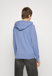 Haut à capuche bleu clair en tissu doux, avec une capuche, des poignets côtelés et un ourlet droit. La vue arrière révèle un design simple sans détails supplémentaires.