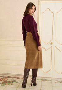 Burgundijas krāsas garajām piedurknēm augšdaļa ar faldu aprocēm, savienota ar brūnu zamša midi svārku un brūnām augstpapēžu zābakām līdz ceļiem. Redzamas gludas tekstūras.