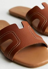 Sandali in pelle marrone con texture e design a intaglio, caratterizzati da cuciture bianche e una suola piatta e liscia. Stile semplice, con punta aperta.