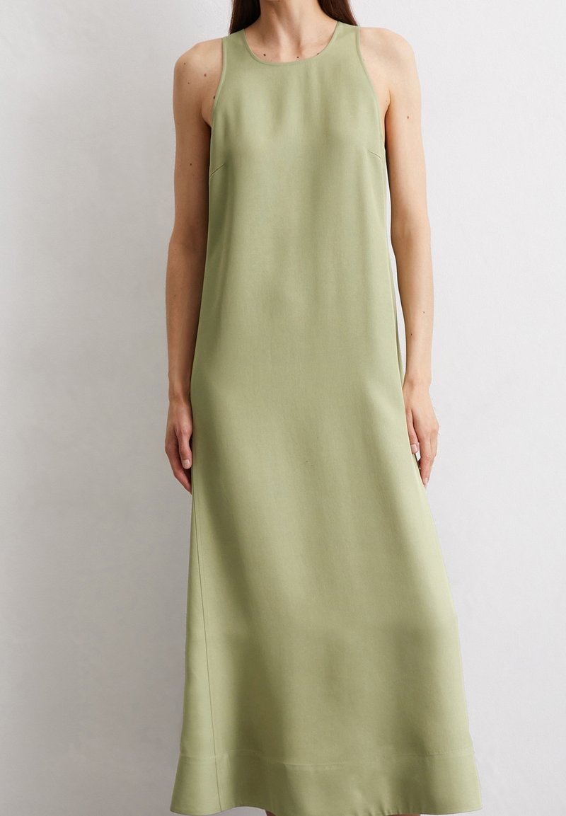 Femme portant une robe sans manches, vert clair, longueur cheville, debout contre un mur uni de couleur claire.
