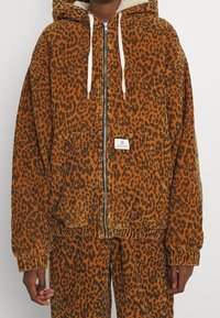 Kamerakuvia näyttävä leopardikuosisella corduroy-takilla, jossa on huppu, etuvetoketju, säätönyörit ja kaksi etutaskua. Raskaastitekstuuri; lämmin oranssi ja musta väri.