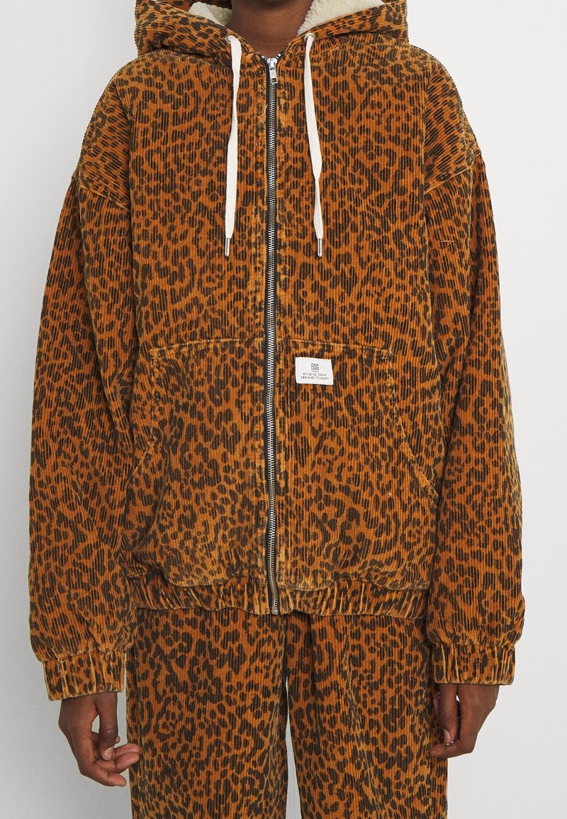 Kamerakuvia näyttävä leopardikuosisella corduroy-takilla, jossa on huppu, etuvetoketju, säätönyörit ja kaksi etutaskua. Raskaastitekstuuri; lämmin oranssi ja musta väri.